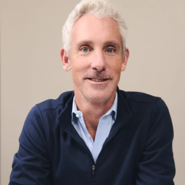 A middle-aged man with short gray hair and light facial hair sits at a table wearing a navy pullover, light blue shirt, and a wristwatch, looking slightly off-camera.