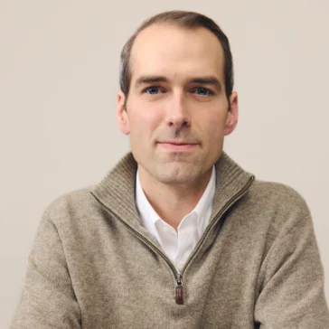 A man in a beige sweater and white shirt sits at a table, looking at the camera and smiling slightly.