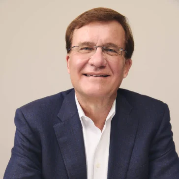 Man wearing glasses, a navy suit jacket, white shirt, wristwatch, and gold ring, sitting at a table with hands folded, smiling at the camera against a neutral background.