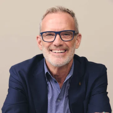 A middle-aged man with gray hair and beard, wearing blue glasses, a navy blazer, and a patterned shirt, sits at a table with arms crossed and smiles at the camera.