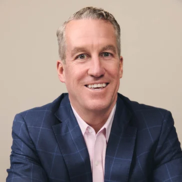 A man in a blue checkered suit jacket, pink shirt, and watch sits at a table with his arms crossed, smiling at the camera against a plain background.