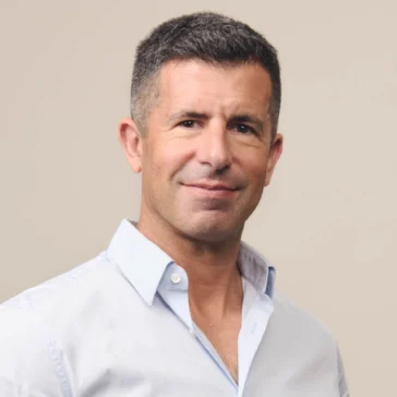 A man with short dark hair wearing a light blue button-up shirt stands against a plain beige background, looking at the camera and smiling slightly.