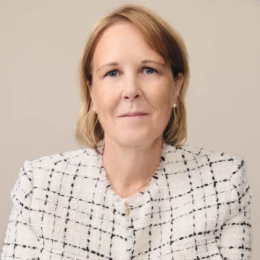 A woman with light brown hair wearing a white and black checkered blazer sits at a table, facing the camera with a neutral expression.