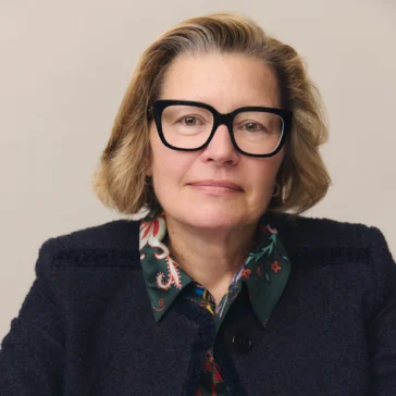 A woman with blonde hair and black glasses sits at a table, wearing a dark blazer over a patterned blouse, with her hands resting in front of her.