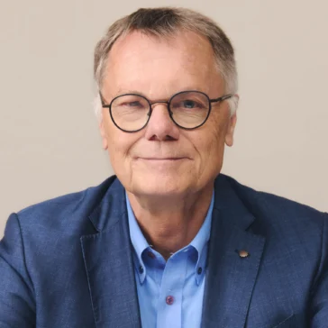 An older man with short gray hair and glasses wears a blue suit jacket over a light blue shirt, sitting with his arms crossed on a table, against a plain background.