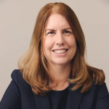A woman with shoulder-length brown hair wearing a dark blazer sits at a table, smiling with her hands folded in front of her against a plain background.