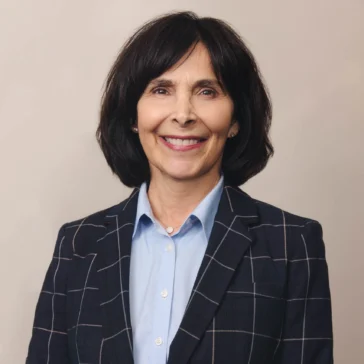 A woman with short dark hair wearing a plaid blazer and light blue shirt stands against a plain background, smiling at the camera.