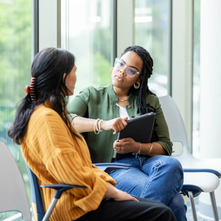 Two women sit beside a window having a conversation; one holds a clipboard and pen, listening attentively while the other speaks.