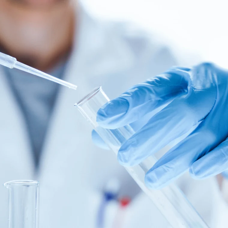 A person wearing blue gloves and a lab coat uses a pipette to transfer liquid into a test tube in a laboratory setting.