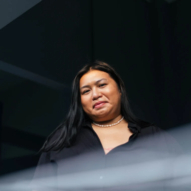 A person with long dark hair and a pearl necklace stands indoors, wearing a black shirt, looking slightly downward with a neutral expression.