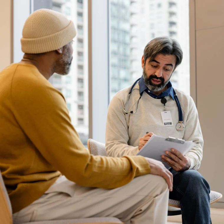 A doctor with a stethoscope and clipboard talks to a patient in a yellow sweater in a modern office with large windows.
