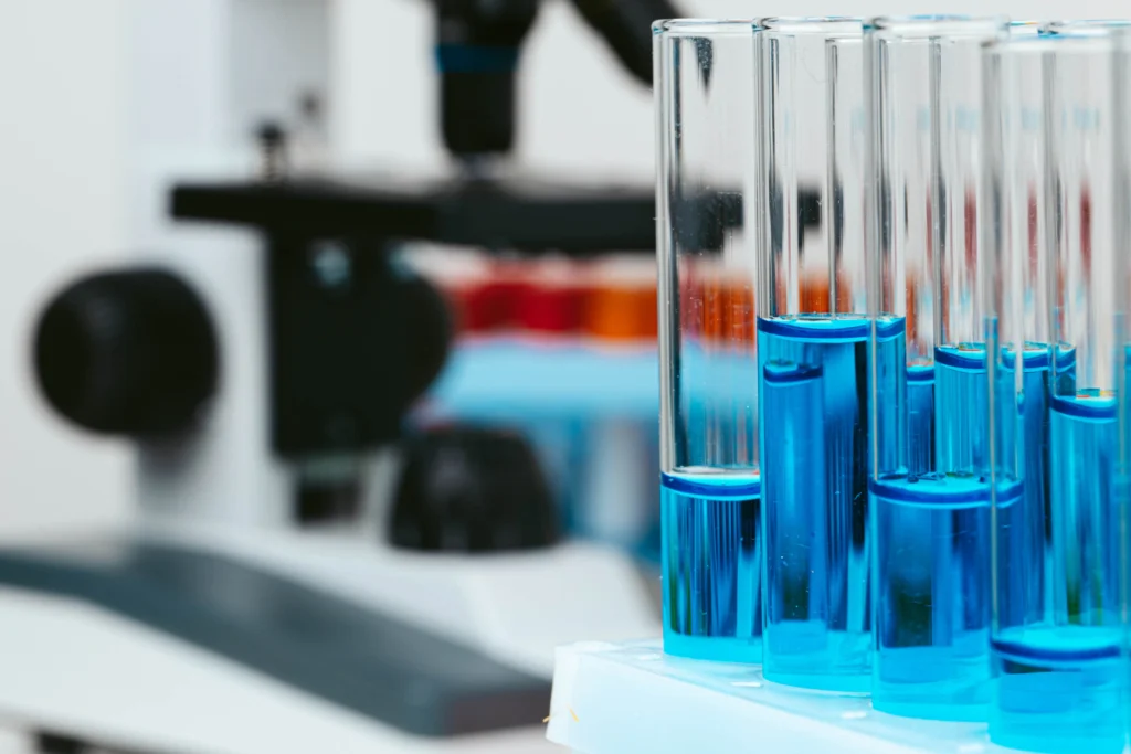 A row of test tubes filled with blue liquid is placed in front of a blurred microscope in a laboratory setting.