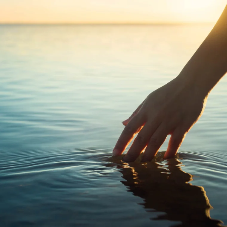 A hand gently touches the surface of calm water at sunset, creating small ripples.