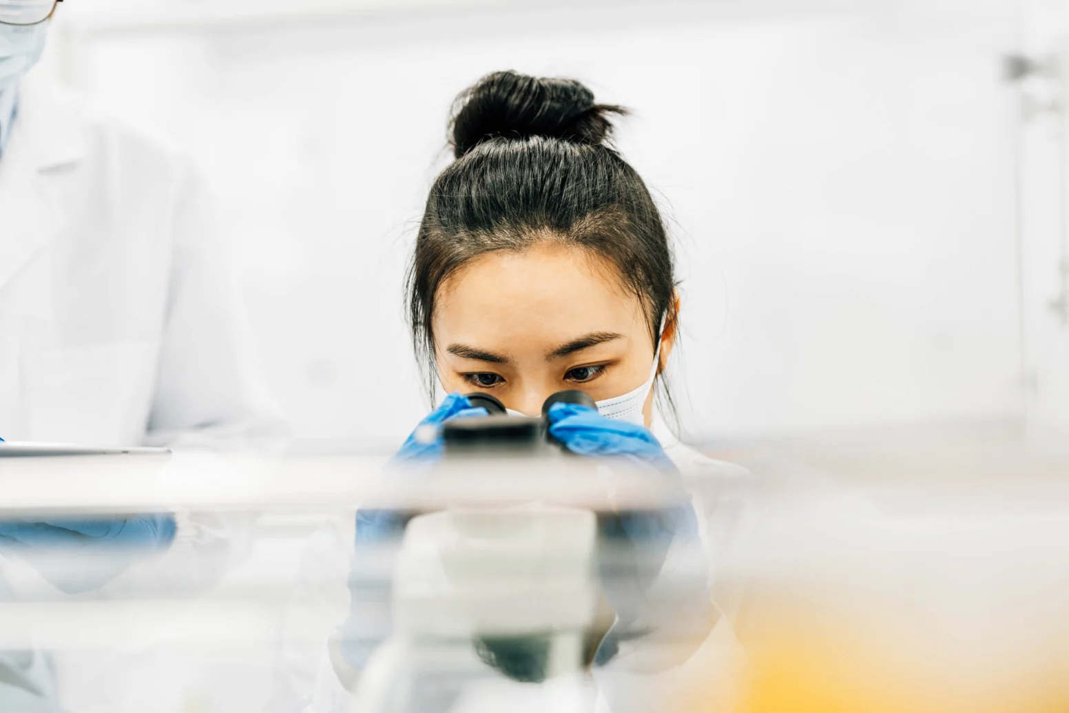 A person in a lab coat and gloves looks through a microscope in a laboratory setting, with another figure partially visible nearby.