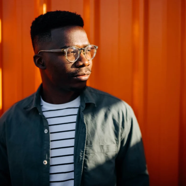 A person wearing glasses, a striped shirt, and an open button-down shirt stands in front of an orange corrugated metal wall, looking to the side.