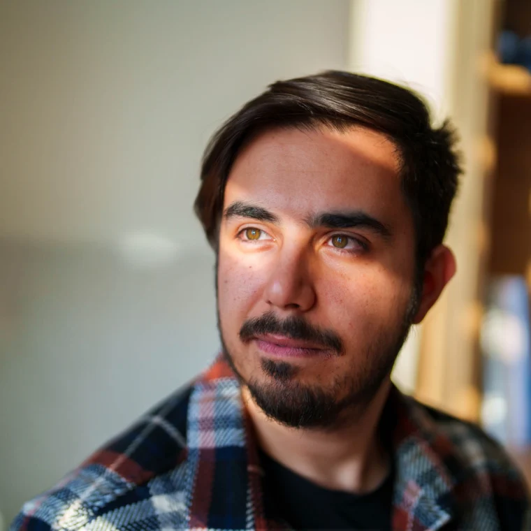 A man with short dark hair and a beard looks to the side, sunlight partially illuminating his face. He is wearing a plaid shirt and appears to be indoors.