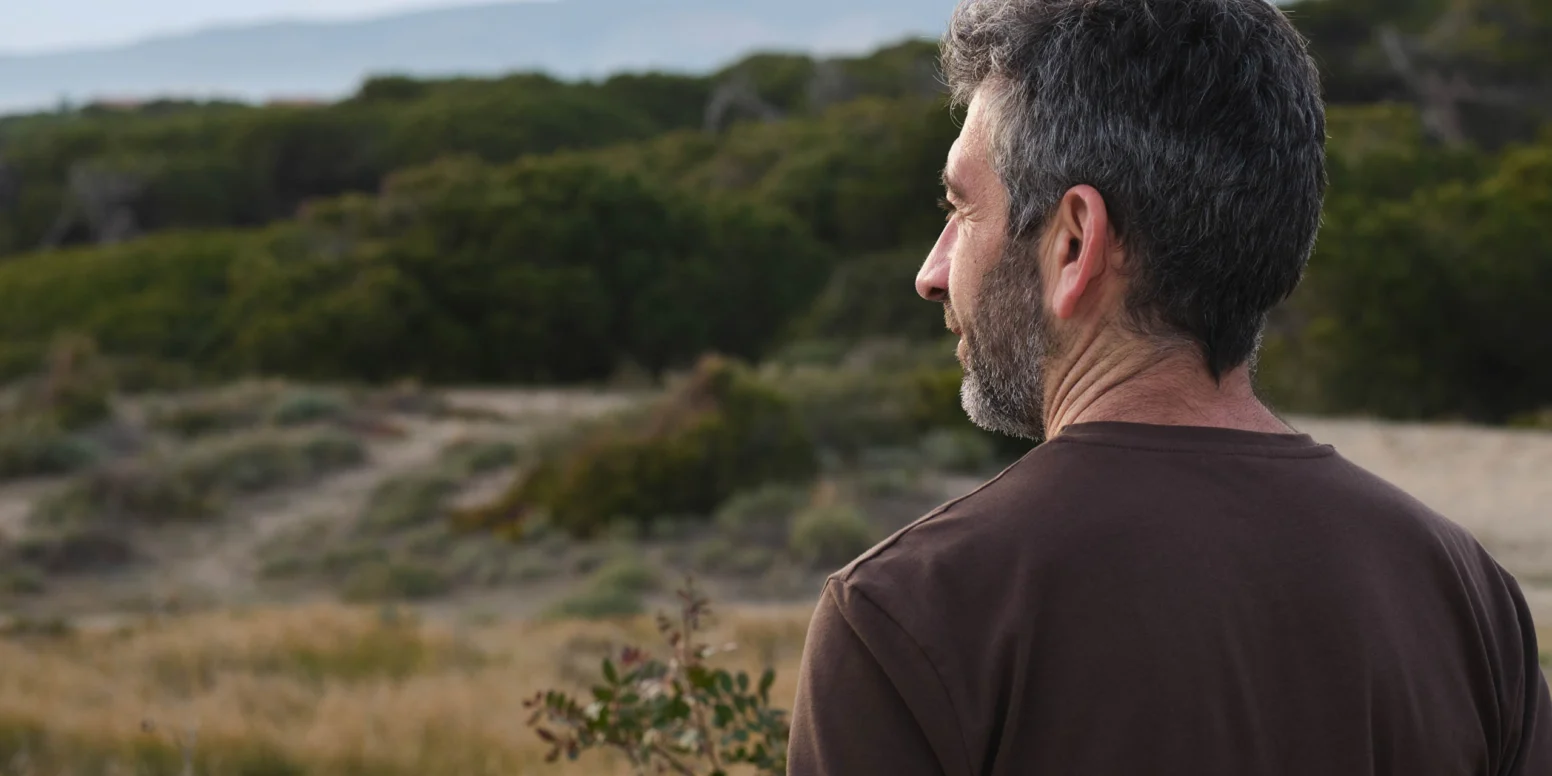 A man with short gray hair and a beard stands outdoors, facing away, looking at a landscape with grass, shrubs, and distant hills.