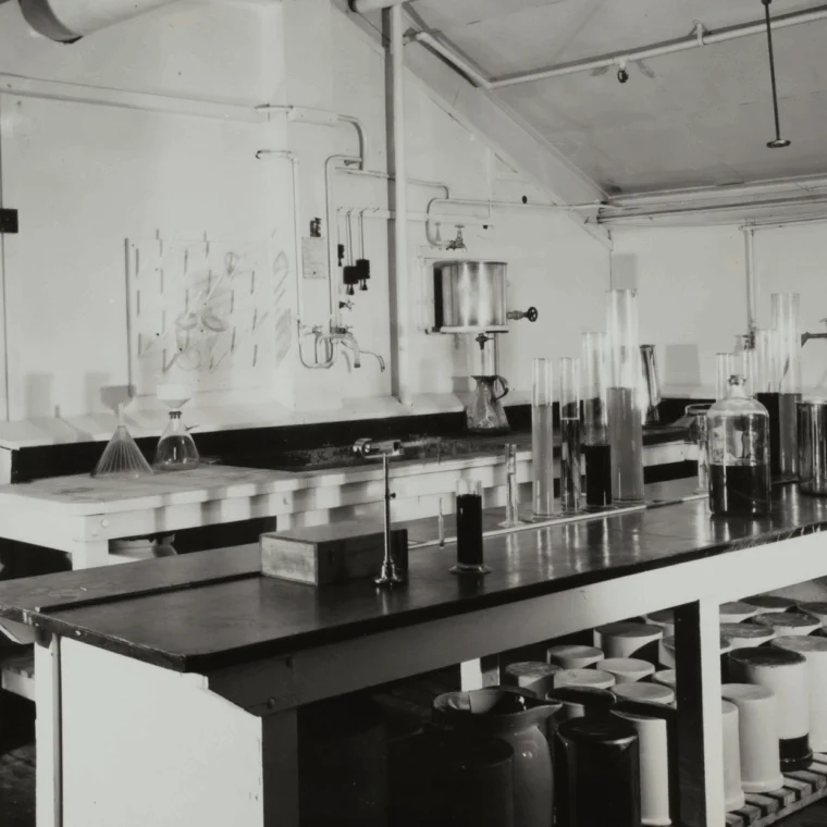 Black-and-white photo of an old laboratory with glassware, bottles, and flasks on countertops, shelves with containers, and pipes along the walls.