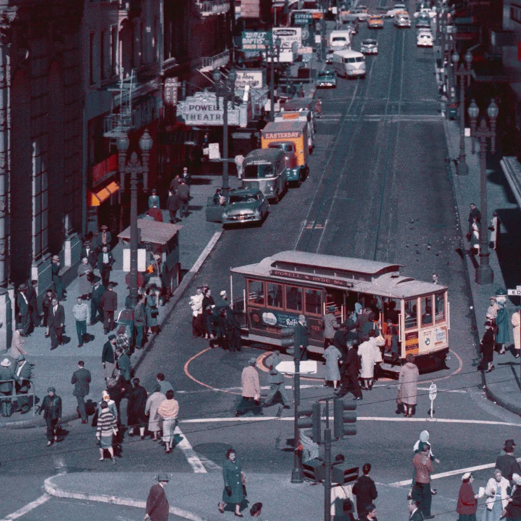 A busy city street scene with a cable car stopped at an intersection while pedestrians cross and cars wait in the background.