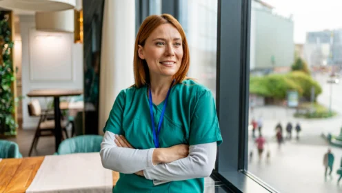 A woman in green medical scrubs stands indoors by a large window, arms crossed, looking outside with a slight smile.