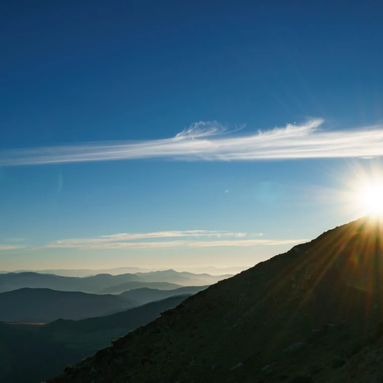 The sun rises over a mountain slope with rays shining, distant mountain ranges, and a clear blue sky with thin clouds.