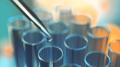 A pipette dispenses a droplet of liquid into one of several glass test tubes, suggesting a laboratory or scientific experiment setting.