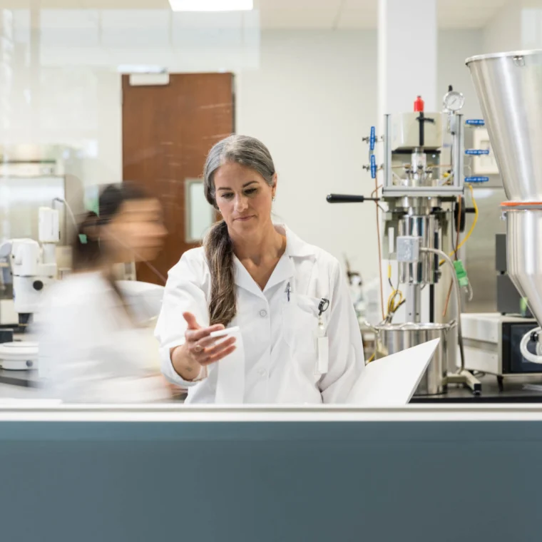 A woman in a lab coat stands in a laboratory, gesturing with her hand, while other people and scientific equipment are visible in the background.