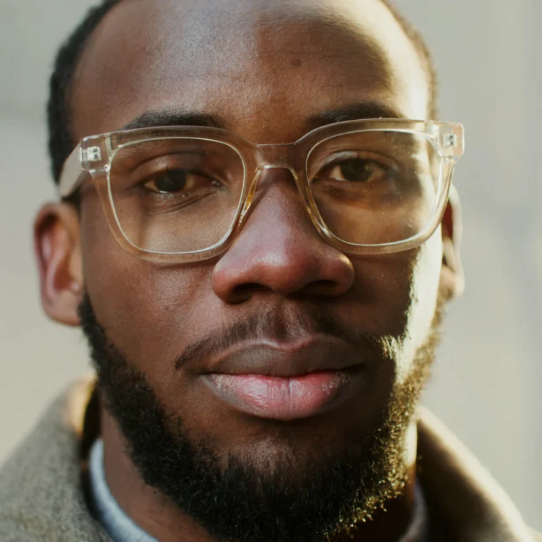 A man with clear glasses, a short beard, and a neutral expression looks directly at the camera. The background is softly blurred.