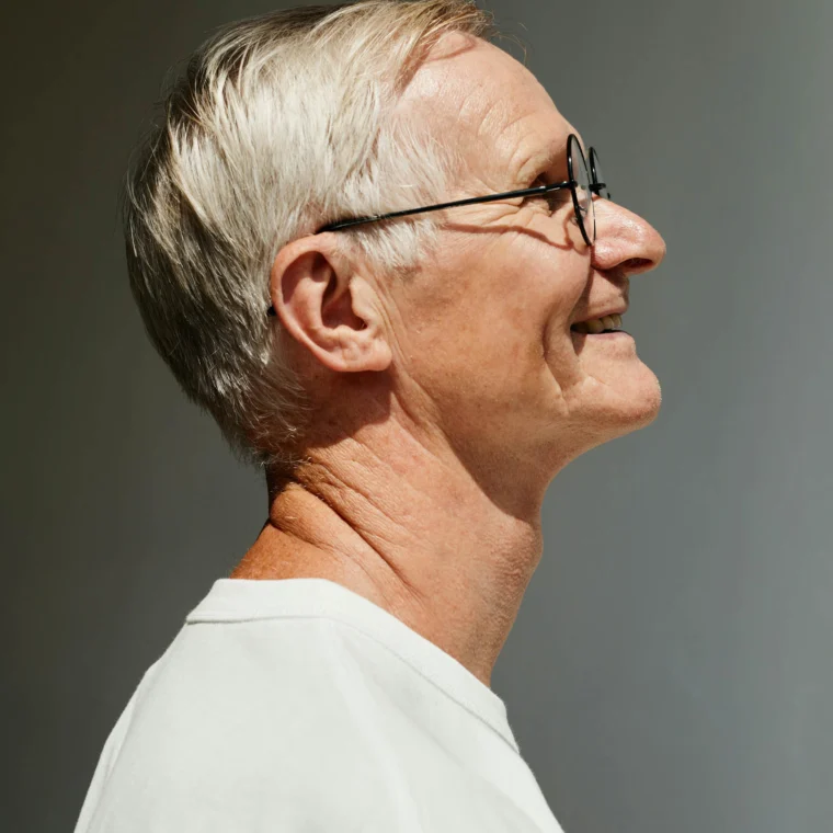 An older man with short gray hair and glasses is smiling, viewed in profile, wearing a white shirt against a neutral background.