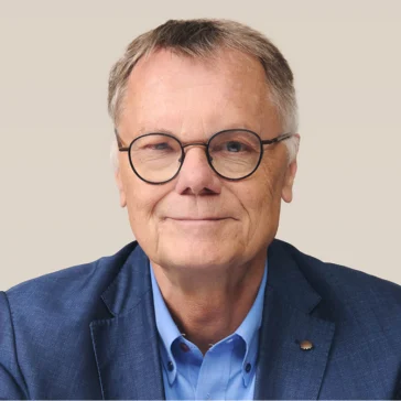 An older man with short gray hair, wearing round glasses, a blue dress shirt, and a blue blazer, sits against a plain light background, looking at the camera.