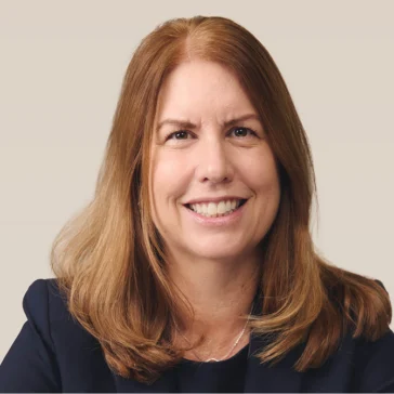A woman with straight, shoulder-length light brown hair wearing a dark blazer is smiling at the camera against a plain light background.