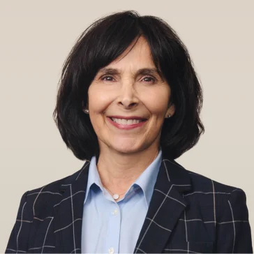 Woman with dark shoulder-length hair wearing a dark plaid blazer and light blue collared shirt, smiling in front of a plain light background.