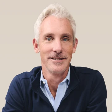 A man with short white hair and light facial hair wearing a navy jacket over a light blue shirt sits against a plain beige background.
