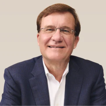 A middle-aged man with short brown hair, glasses, and a blue blazer over a white shirt, smiling and sitting against a plain light background.