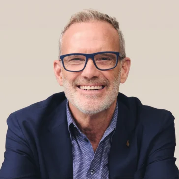 A middle-aged man with short gray hair, a trimmed beard, and blue glasses is smiling, wearing a blue blazer and patterned shirt against a plain light background.