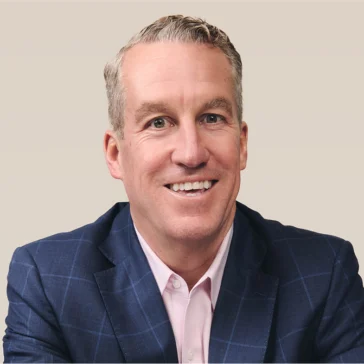 A man in a blue checkered suit jacket and light pink shirt is smiling, sitting against a plain beige background.