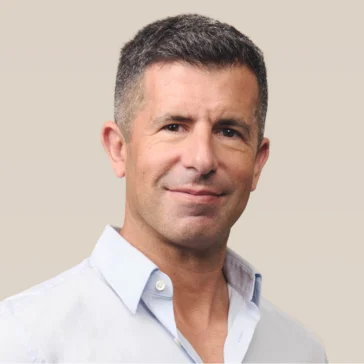 A man with short dark hair and a trimmed beard wears a light blue collared shirt and smiles at the camera against a plain beige background.