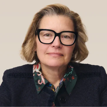A middle-aged woman with short blonde hair, wearing large black glasses, a dark textured jacket, and a green patterned scarf, looks directly at the camera against a plain background.