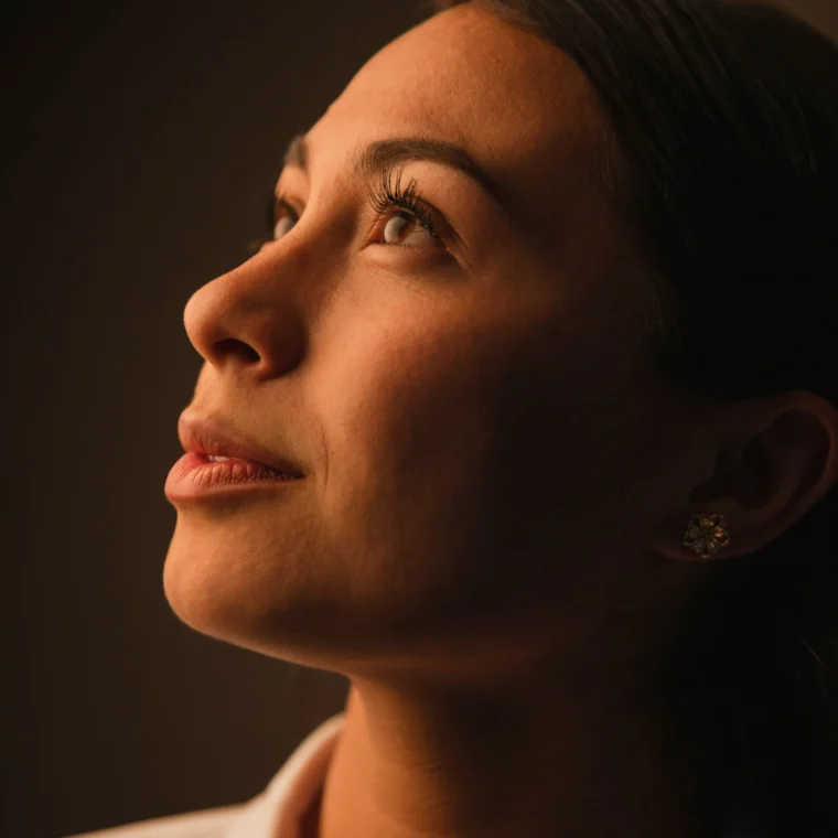 A woman with long dark hair looks upward, illuminated by warm light against a dark background.