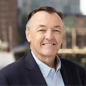 Middle-aged man with short brown hair wearing a navy blazer and light blue shirt, smiling in an outdoor urban setting with blurred buildings in the background.