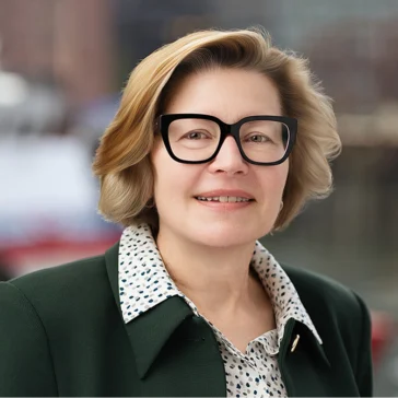 A woman with short blonde hair, wearing black glasses, a white patterned blouse, and a dark green blazer, smiles at the camera with a blurred cityscape in the background.