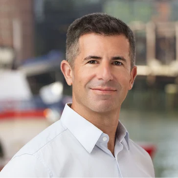 Man with short dark hair and light blue dress shirt standing outdoors, blurred background with water and boats.