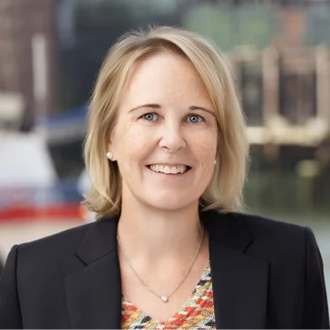 A woman with shoulder-length blonde hair, wearing a navy blazer and patterned top, smiles at the camera with a blurred outdoor background.