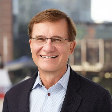 A man with short brown hair and glasses, wearing a dark blazer and light blue shirt, smiles at the camera with a blurred cityscape in the background.