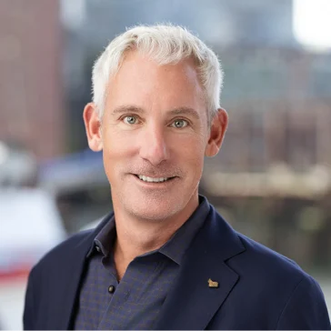 A man with short white hair wearing a navy blazer and patterned shirt smiles at the camera, with an urban waterfront blurred in the background.
