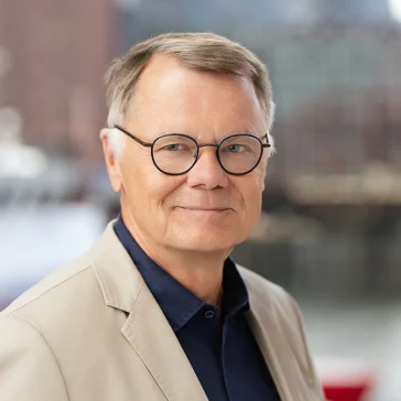 Older man with short light hair, glasses, and a beige blazer over a dark shirt, standing outdoors with a blurred urban waterfront background.