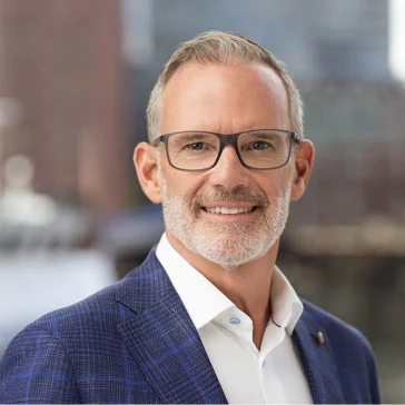 Smiling middle-aged man with short gray hair, beard, and glasses, wearing a blue plaid suit jacket and white shirt, standing in front of a blurred urban background.