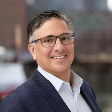 Smiling man in glasses and a blue checkered suit jacket, standing outdoors with a blurred urban background.