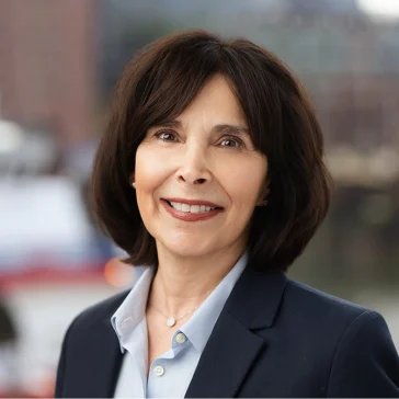 Woman with shoulder-length brown hair wearing a navy blazer and light blue shirt, smiling outdoors with an urban background.