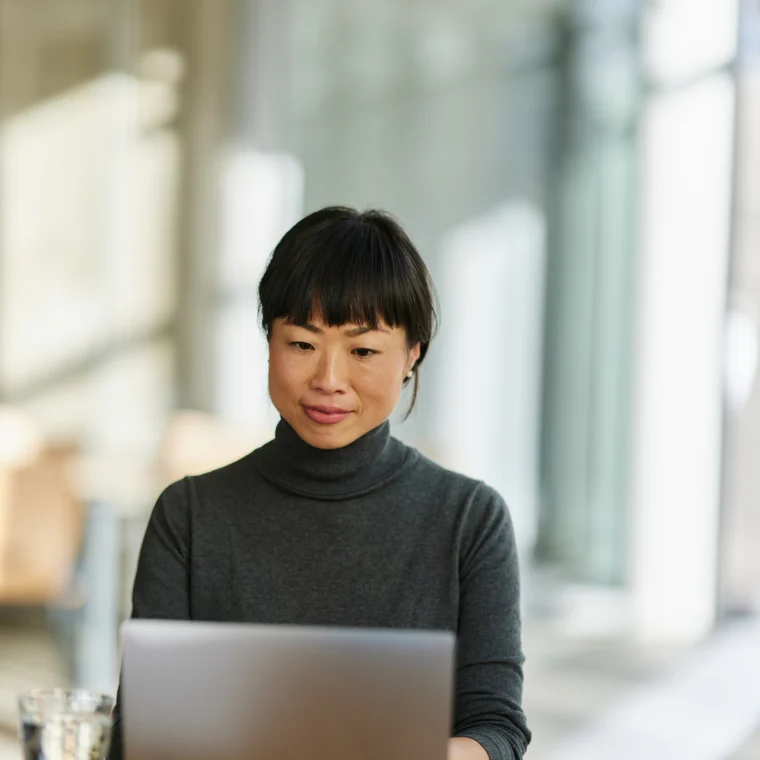 A person with short black hair in a gray turtleneck sits at a table working on a laptop in a bright, modern indoor space. A glass of water and papers are on the table.